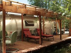 an airstream sits on a wooden deck next to some chairs and tables in the shade