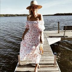 a woman wearing a hat and dress walking on a dock near the water with her hand in her pocket