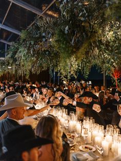 a large group of people sitting at tables with candles in front of them and trees