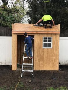 two men are working on the roof of a small wooden shed while another man stands on a ladder