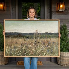 a woman holding up a large painting in front of a house with potted plants