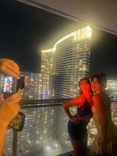 two women taking pictures with their cell phones in front of a cityscape at night