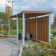 two people are sitting on a bench under a wooden structure in the middle of a park