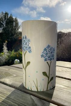 a white coffee cup with blue flowers painted on it sitting on a wooden table outside