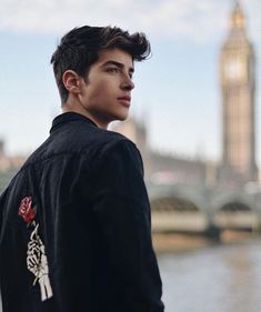 a young man standing in front of a river with a clock tower in the background