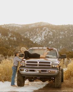 two people standing next to a truck in the middle of a field with mountains in the background