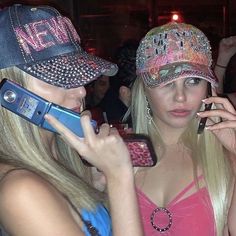 two beautiful young women standing next to each other while talking on cell phones and wearing hats