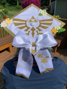 A white graduation cap with a sparkly gold boarder. The hat has yellow and white flowers as well as a sparkling gold triforce symbol. There is a white bow fixed to the bottom corner with the initials SVG  and the words “Drexel 24” on one tail of the bow. The other tail has an image of a silent princess in the same sparkling gold as the rest of the hats detail. Splatoon Graduation Cap, Grad Cap Studio Ghibli, Jjba Graduation Cap, Gojo Graduation Cap, Graduation Cap Designs Coraline, Legend Of Zelda Graduation Cap, Undertale Graduation Cap