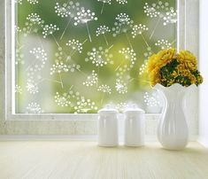 two white vases with yellow flowers sit in front of a window that has dandelions on it