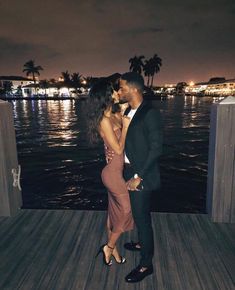 a man and woman kissing on a dock by the water at night with palm trees in the background