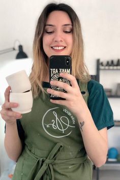 a woman in an apron is taking a selfie with her cell phone and cup