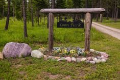 a wooden sign sitting in the middle of a forest next to a dirt road and trees