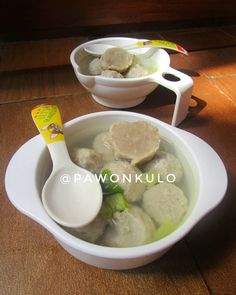 two white bowls filled with food on top of a wooden table next to spoons