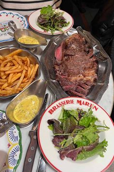 a table topped with plates and bowls filled with meat next to french fries, salad and sauces