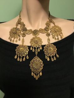 For sale is a stunning antique brass necklace from the Victorian era, featuring a beautiful filigree bib design in Etruscan Revival style. The 6 filigree pendants are round with a diameter of 1.5" and they are adorned with dangles, while the necklace itself is 14" in inner circumference with a 6.5" drop. The necklace is pre-owned with a box closure. As can be seen in photo #7, there is minor corrosion on one medallion, but otherwise, the necklace is in very good condition. This unique vintage piece is a statement accessory that will add a touch of sophistication to any outfit. The brass/gold color complements the intricate metalwork, and the absence of any stones allows the design to speak for itself. This necklace is a true reflection of original Victorian fashion and a must-have for any Elegant Festival Necklace With Intricate Design, Bohemian Gold Jewelry With Intricate Design, Antique Necklaces With Vintage Charm For Ceremonial Occasions, Elegant Filigree Necklaces For Festivals, Traditional Gold Necklaces With Vintage Charm, Gold Brass Necklace With Antique Finish, Antique Brass Necklaces For Ceremonial Occasions, Antique Brass Necklace For Ceremonial Occasions, Metal Filigree Jewelry For Vintage Style