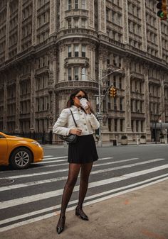 a woman standing in front of a tall building drinking from a coffee cup while wearing black tights and high heels