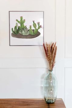 a glass vase with some plants in it on a wooden table next to a painting