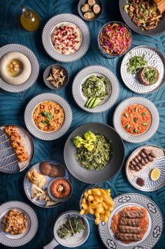 an overhead view of several plates of food on a blue tablecloth with silverware and utensils