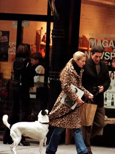 a man and woman walking their dog down the street in front of a magazine store