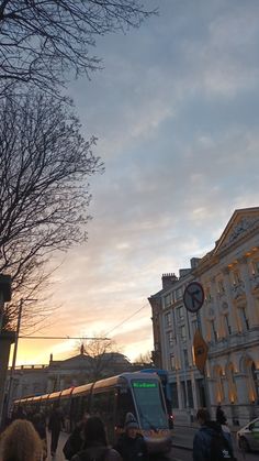 people are standing on the side walk near a bus and some buildings at sunset or dawn