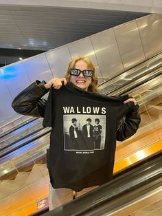 a woman holding up a t - shirt with the words wallows on it in front of an escalator