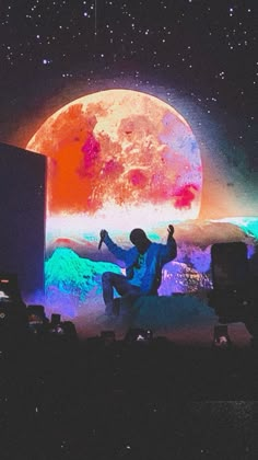 a man sitting on top of a stage in front of a giant moon with his hands up