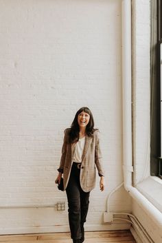 a woman standing in front of a window smiling at the camera while wearing a blazer and black pants