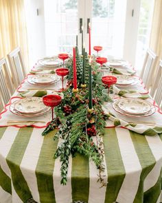 the table is set for christmas dinner with green and white striped linens, red candlesticks, greenery, and silverware