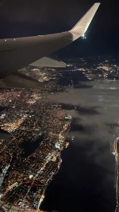 the wing of an airplane flying over a city at night with lights and clouds in the foreground