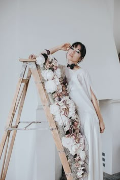 a woman in a white dress standing next to a ladder with flowers all over it