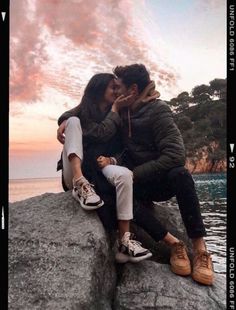 two people sitting on top of a rock next to the ocean with clouds in the background