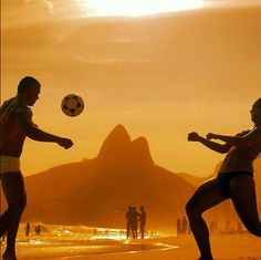 two men are playing soccer on the beach at sunset with mountains in the back ground