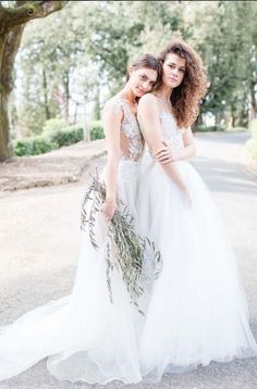 two women in wedding gowns standing next to each other on the road with trees behind them