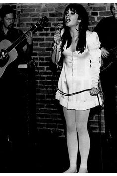 a woman standing in front of a brick wall holding a guitar and singing into a microphone