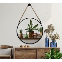 a round mirror hanging on the wall above a wooden dresser with plants and vases