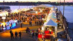 people are walking around an outdoor market by the water at night with lights on them