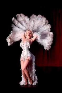 a woman in a white feathered costume is posing on stage with her arms behind her head