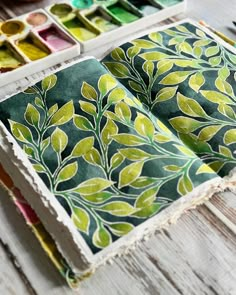 an open book sitting on top of a wooden table next to paintbrushes and paints