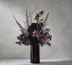 a black vase filled with lots of purple flowers on top of a white table next to a gray wall