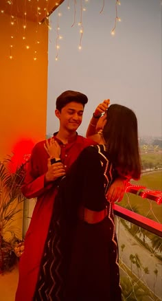 a man and woman standing next to each other on top of a balcony with string lights