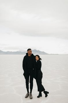 a man and woman standing in the middle of an empty field with mountains in the background