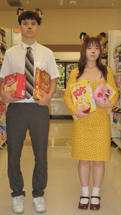 two people standing in a grocery store holding cereal boxes and candy bar bags, one person is dressed as a cat