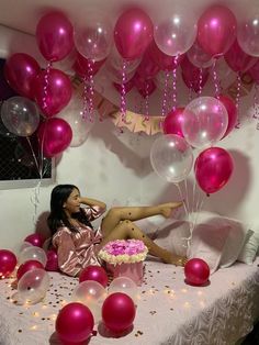 a woman laying on top of a bed surrounded by pink and white balloons, with a cake in front of her