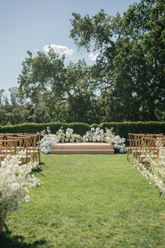 an outdoor ceremony set up with wooden chairs and white flowers on the grass in front of trees