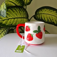 a red and white coffee mug with strawberries on it next to a green plant