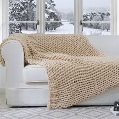 a white couch with a blanket on it in front of a window and snow covered trees outside