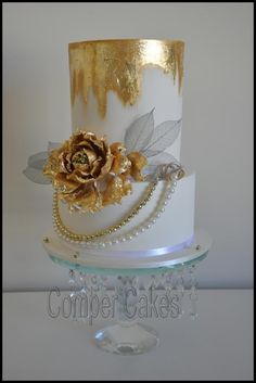 a white and gold wedding cake with flowers on the top is sitting on a glass stand