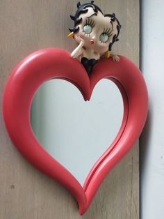 a red heart shaped mirror with a doll on it's head in the shape of a heart