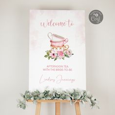 a welcome sign with pink flowers and a tea cup on it, sitting on an easel