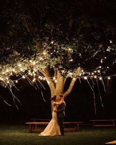 a bride and groom standing under a tree with fairy lights strung all over it at night